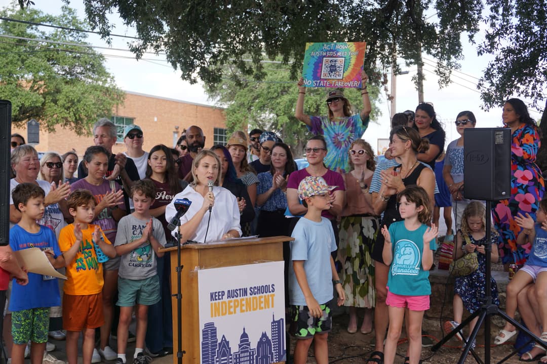 A large group of parents, educators, community members and children gather outside of a school, under a tree, in front of a podium that reads "Keep Austin Schools Independent." A supporter holds a tyedye sign with a QR code prompting readers to sign a petition. Lily McClure speaks at the microphone while supports clap and cheer. 