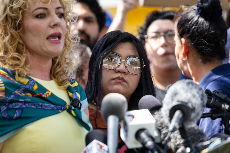 A woman with dark hair and large glasses listens to someone talk at a podium full of microphones.