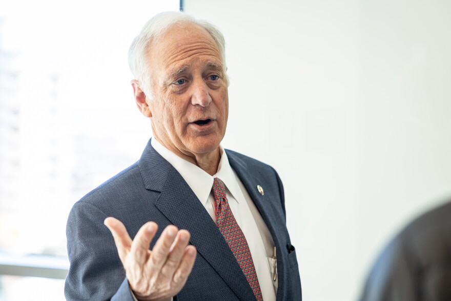 A man in a suit and tie speaks while gesturing with one arm.