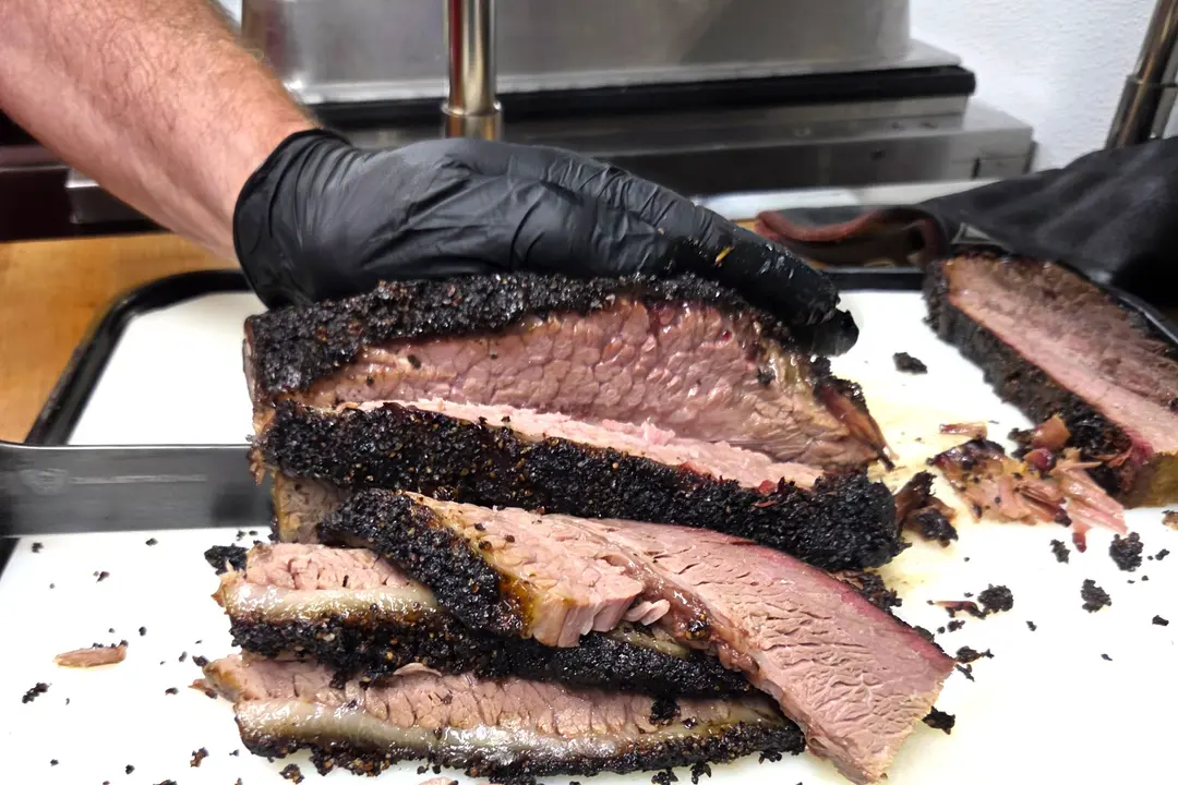 Gloved hand slices smoked brisket with dark peppery bark and pink smoke ring on a cutting board.