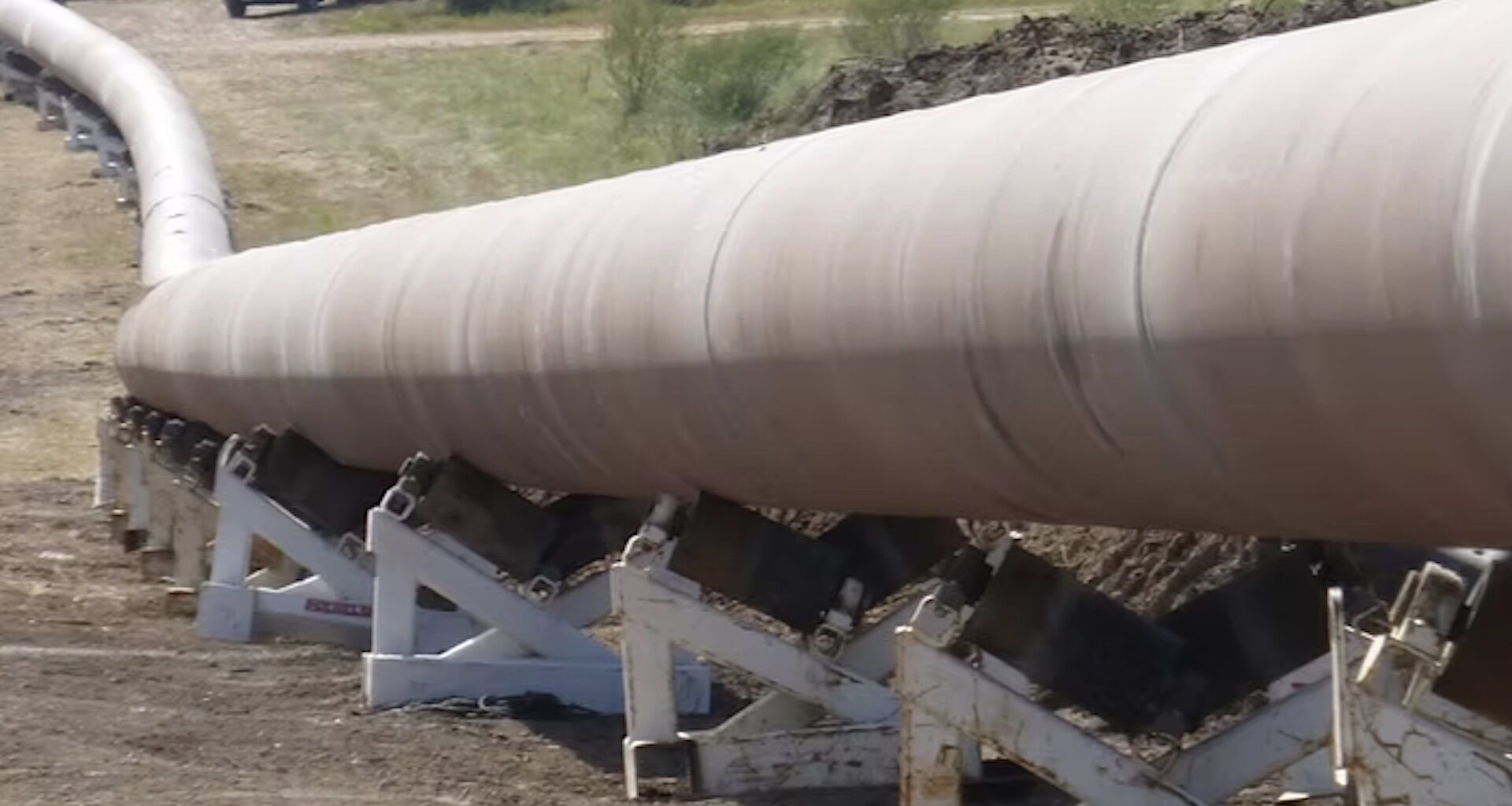 A long pipeline supported by white brackets stretches across a dirt landscape.