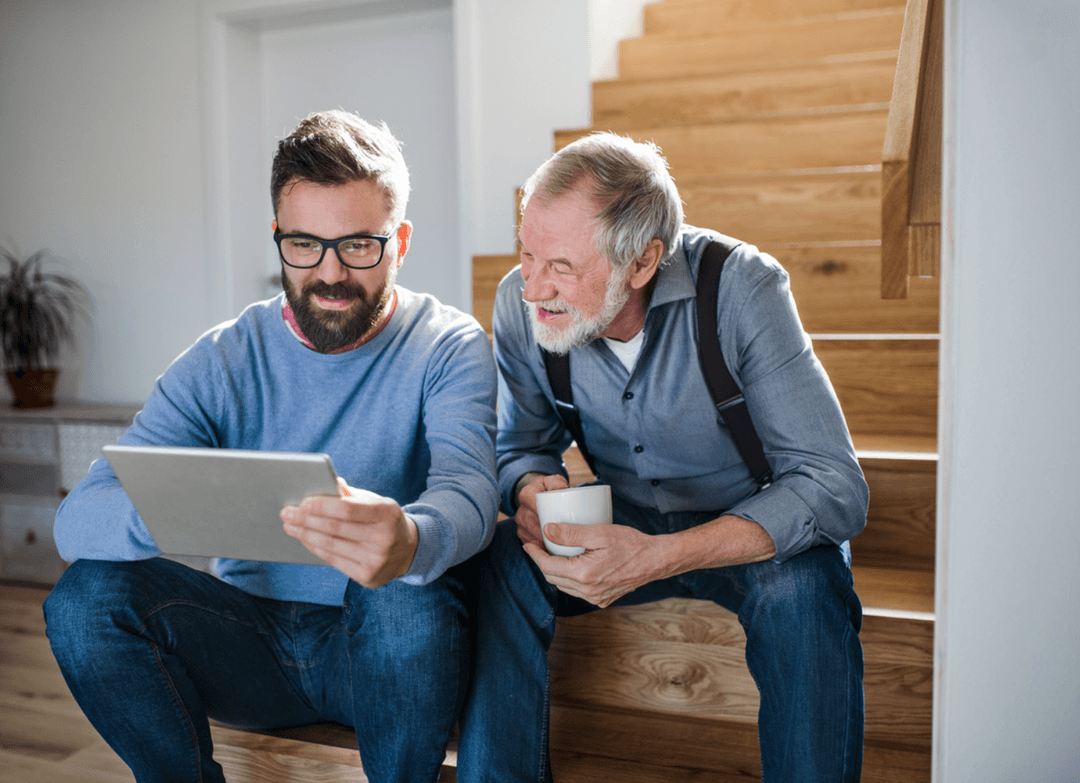 A younger man shows a tablet to a smiling older man holding a mug as they sit on wooden stairs.