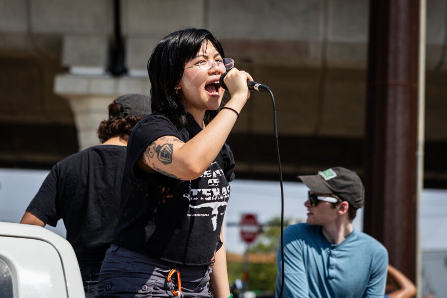 A woman with black hair and a black T-shirt yells into a microphone while standing in the bed of a truck on a sunny day.