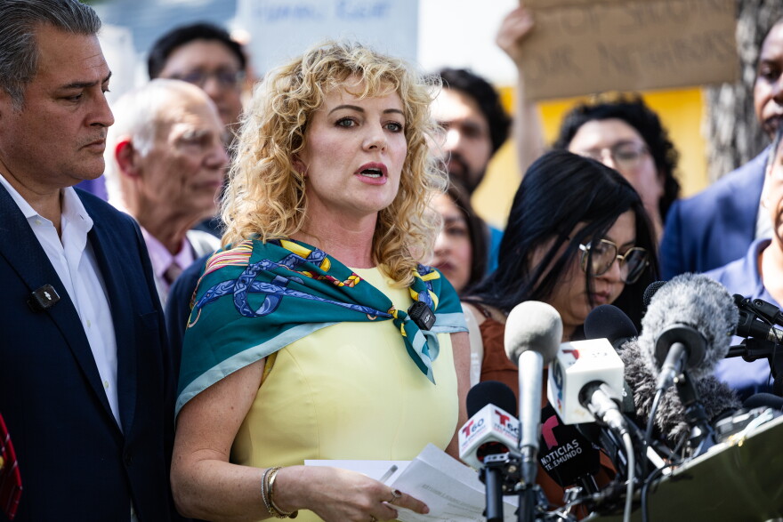A woman, surrounded by people, speaks towards a podium covered in microphones.