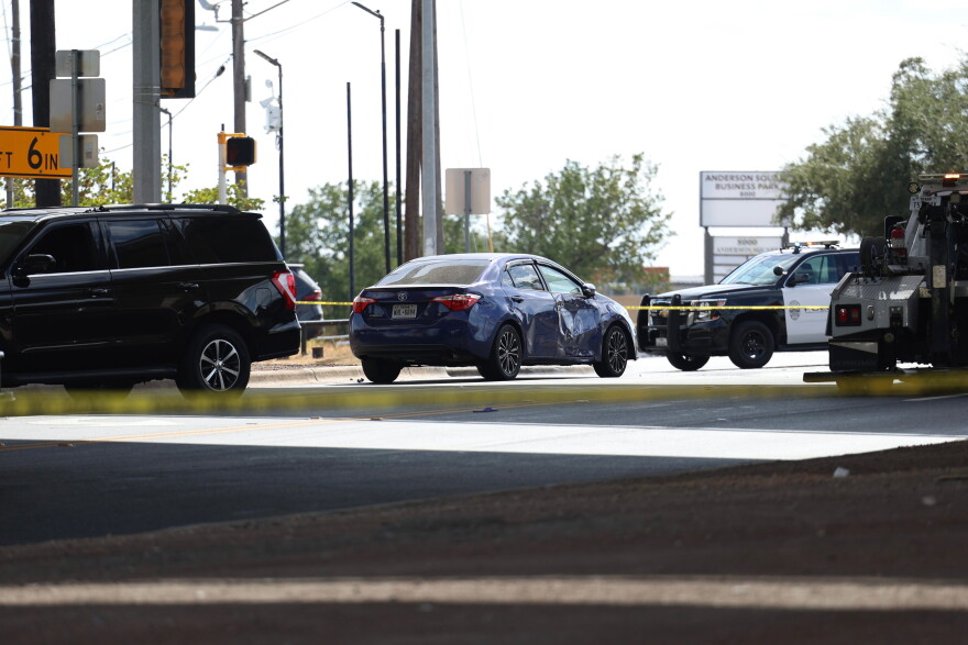 A blue care with a damaged passenger door surrounded by police tape. A police car and a tow truck are nearby.