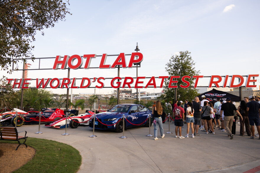 A crowd forms a line in front of the Hot Lap ride. Several cars are parked at the entrance.