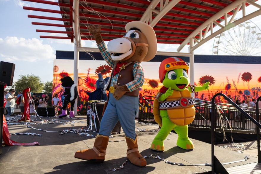 A horse mascot and turtle mascot wave to people from a stage.