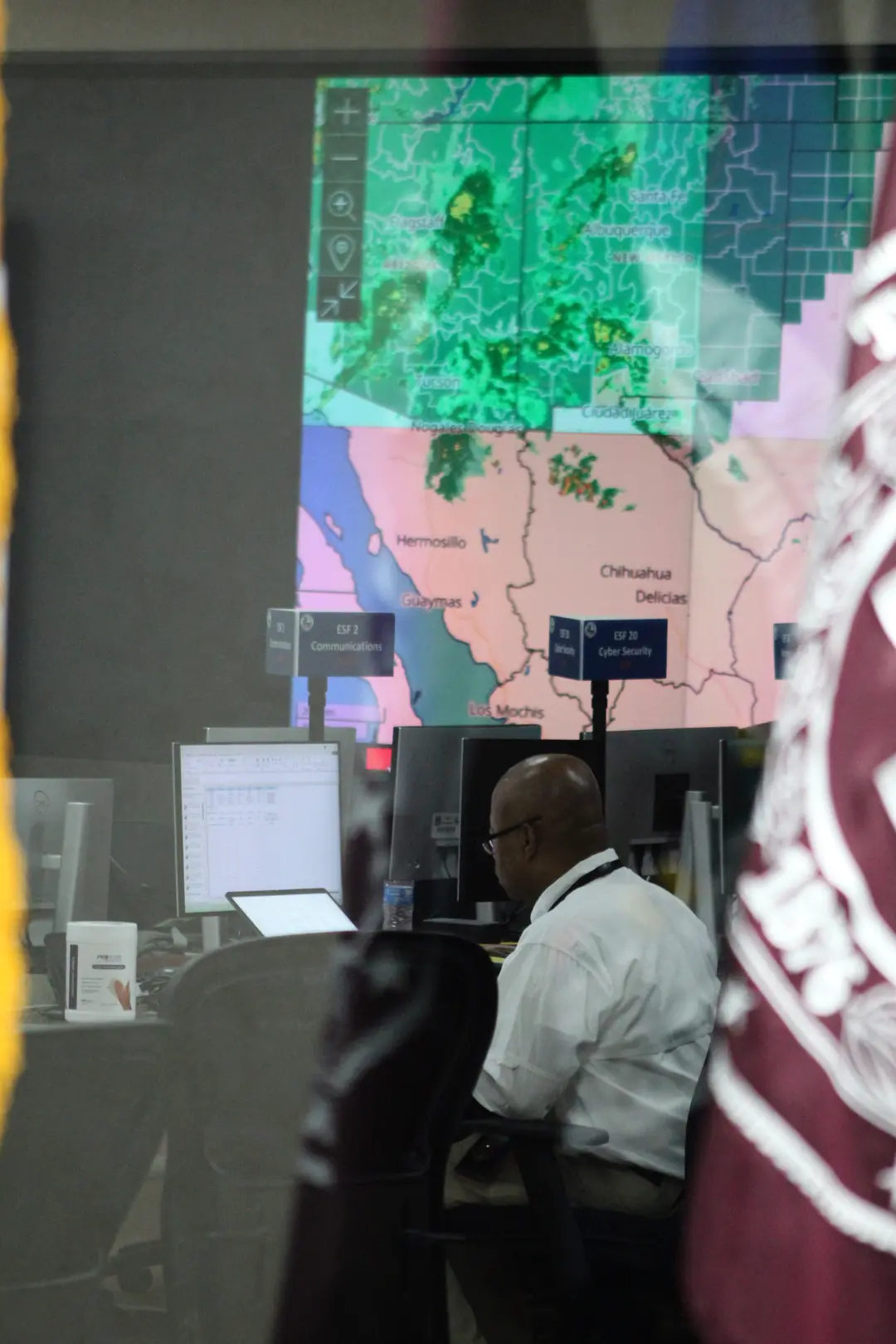 A man in a white shirt sits at a desk, with a weather map displayed on a large screen in the background.