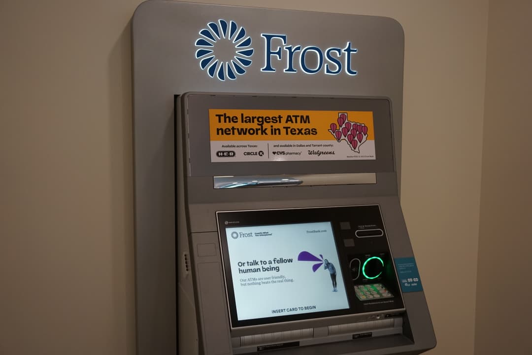 A silver ATM with an illuminated Frost logo on top. A yellow-orange sticker reads "The largest ATM network in Texas," listing H-E-B and Circle K as locations across Texas and CVS and Walgreens as available in Dallas and Tarrant county. The screen reads "Or talk to a fellow human being" before stating "Our ATMs are user friendly, but nothing beats the real thing." The bottom of the screen prompts users to "insert card to begin."