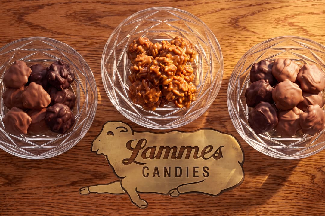 Three glass bowls of candy on a wooden table: milk and dark chocolate-covered pecan candies on either side and caramel pecan pralines in the center. A brass Lammes Candies lamb logo is inlaid in the wood below them.