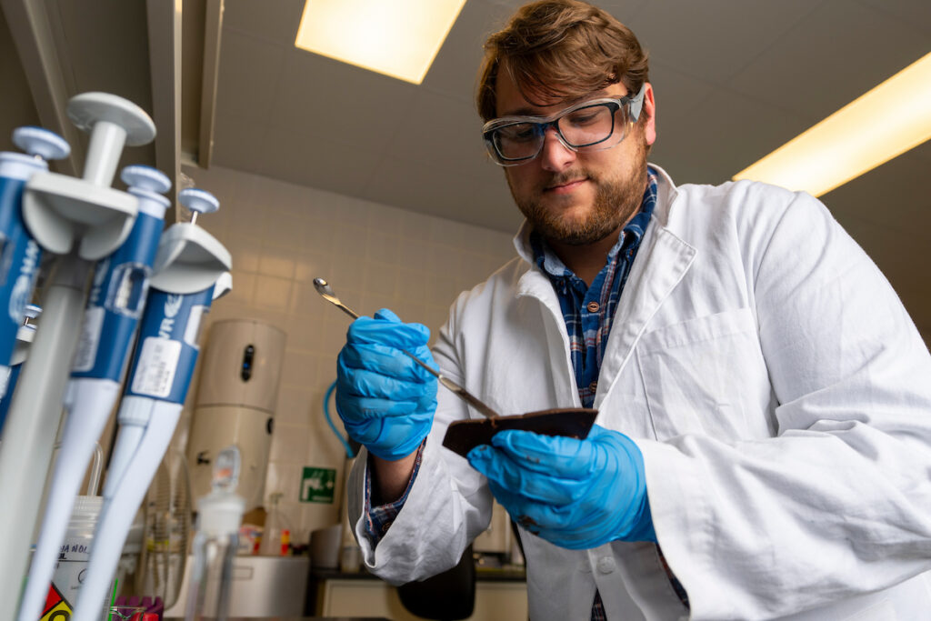 A man in a white coat in a lab looking at lab tools in his gloved hands