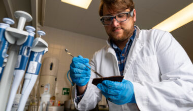 A man in a white coat in a lab looking at lab tools in his gloved hands