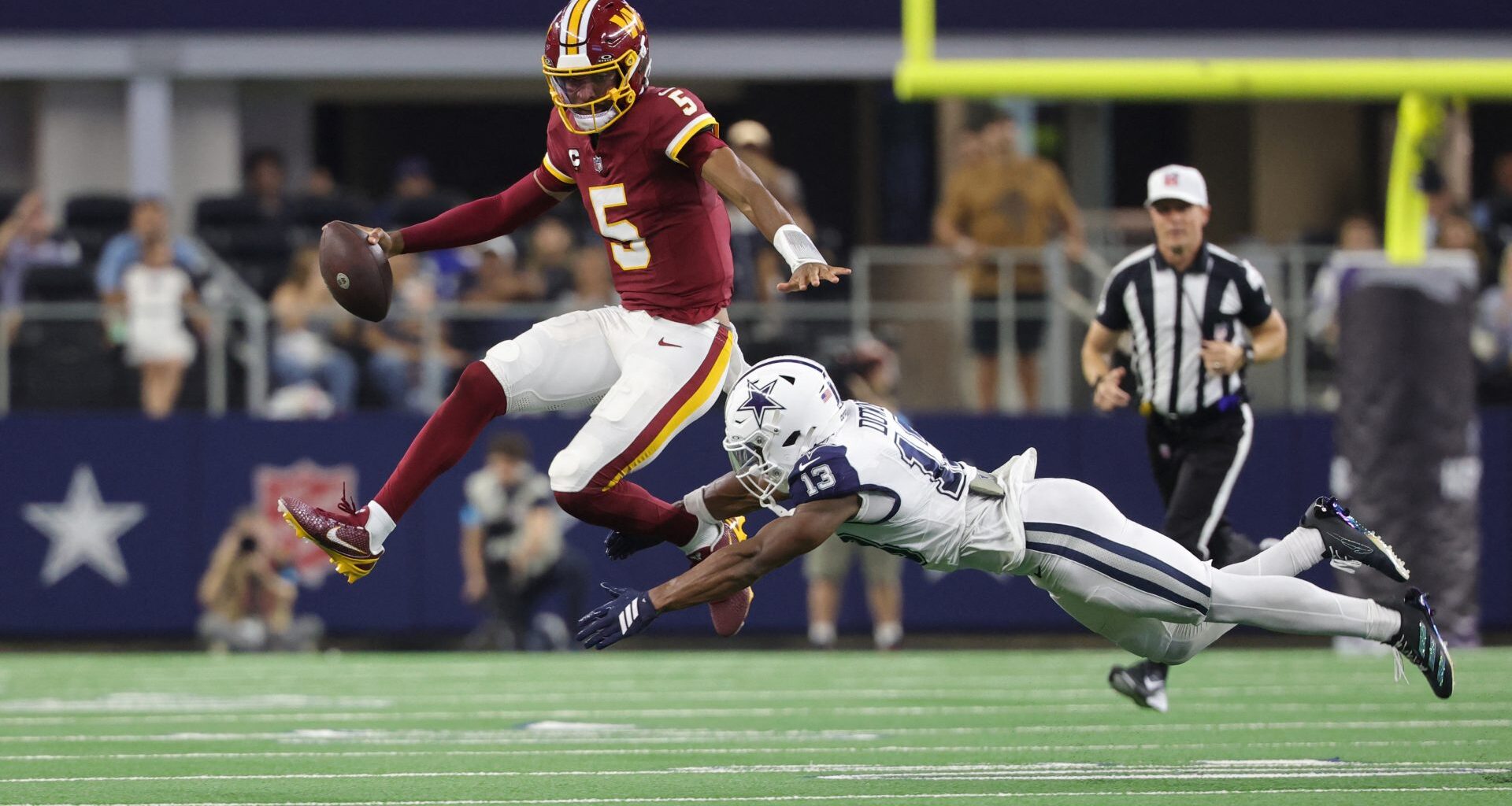 Sep 20, 2026; Arlington, Texas, USA; Washington Commanders quarterback Jayden Daniels (5) leaps to avoid a tackle by Dallas Cowboys safety Caleb Downs (13) while running with the ball during the second half at AT&T Stadium.