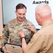 NAMRU San Antonio Director awarded Navy and Marine Corps Commendation Medal for Meritorious Service A military awards ceremony is taking place where a person in a tan/khaki uniform is pinning a medal onto a soldier in Army OCP (Operational Camouflage Pattern) uniform with the name tape 'DIXON'. The soldier is holding a Department of the Navy certificate identifying the award as the Navy and Marine Corps Commendation Medal. The word 'Awards' is visible on a presentation screen in the background.