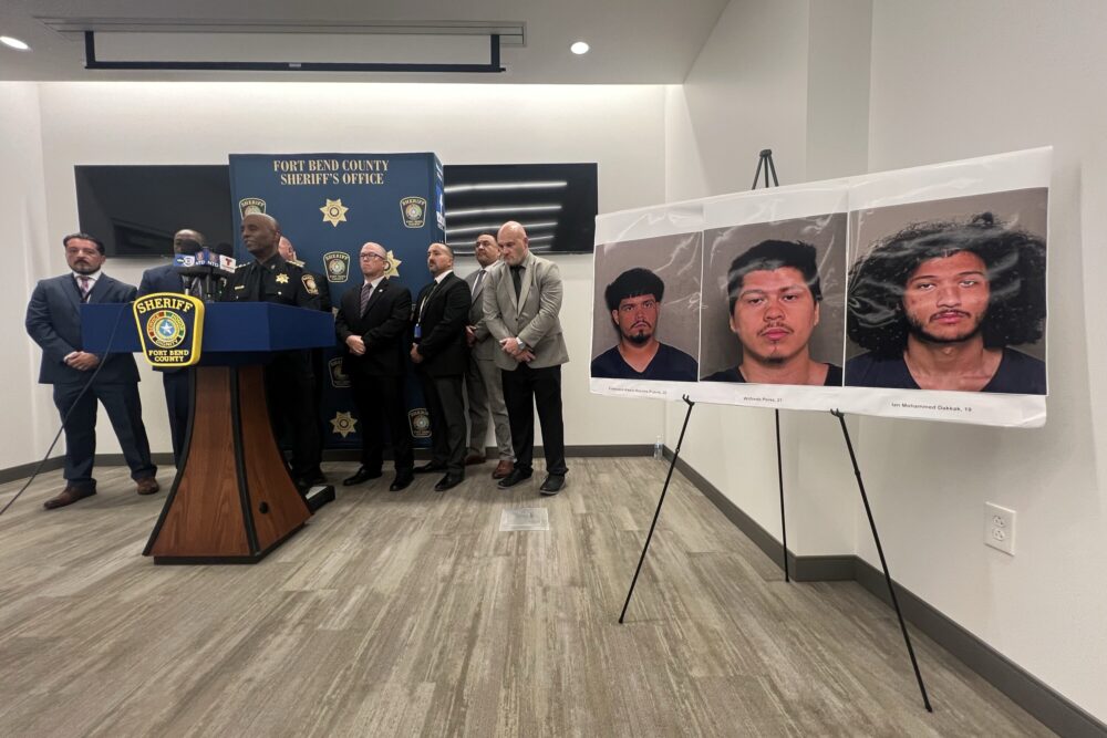 Law enforcement officials standing behind a podium next to an enlarged poster of three suspects identified.