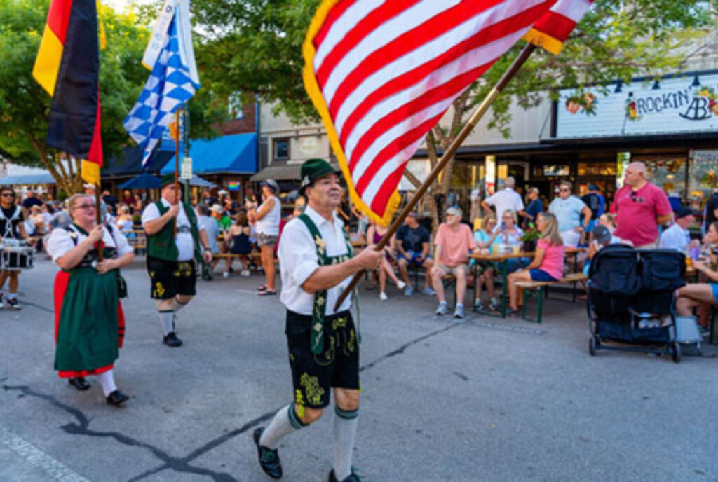 Image shows a man dressed in traditional German attire holding up an American flag.