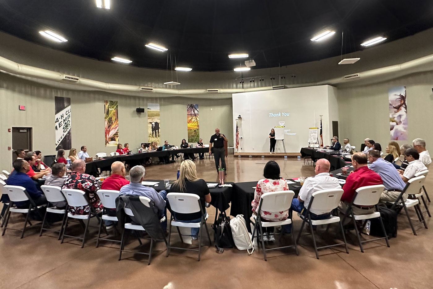 Participants sit around U-shaped tables while Williams speaks during a meeting in a large conference room.