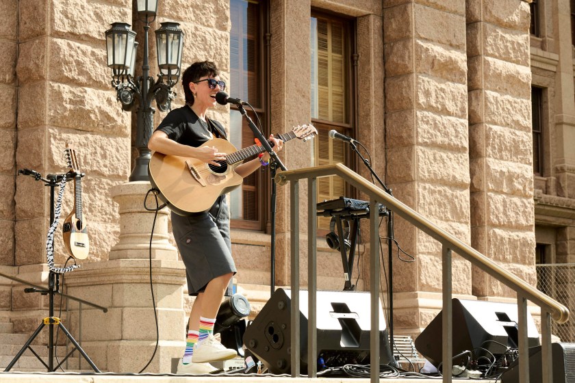 A person in a black T-shirt plays a guitar on the steps of a state building on a sunny day.