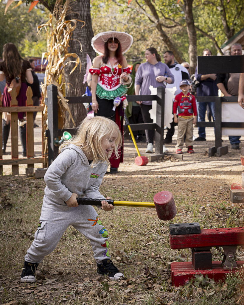 A young child with blonde hair, wearing a gray hoodie and pants, is swinging a mallet at a pumpkin-themed game during a fall festival, with adults and children in the background.