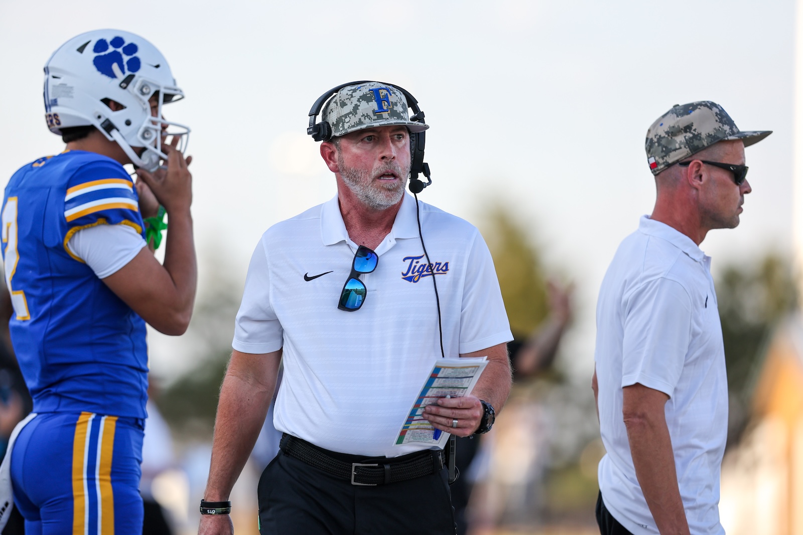 Seabolt Chase-16897 | Press Pass Sports Football coach wearing a headset and sunglasses on the sideline, holding a play sheet near a player in a blue and gold uniform.