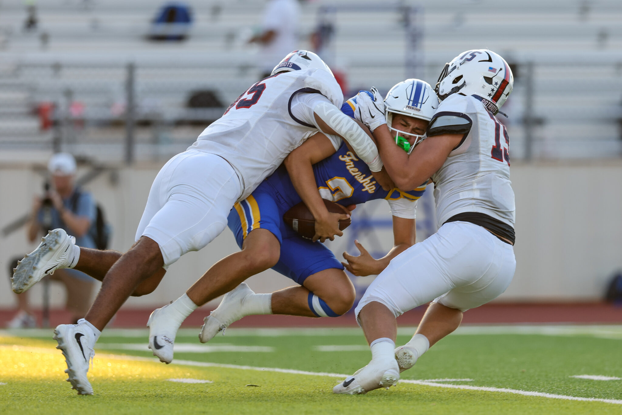 Football players in white tackle a blue-and-gold opponent during a game on a green field with stands in the background.
