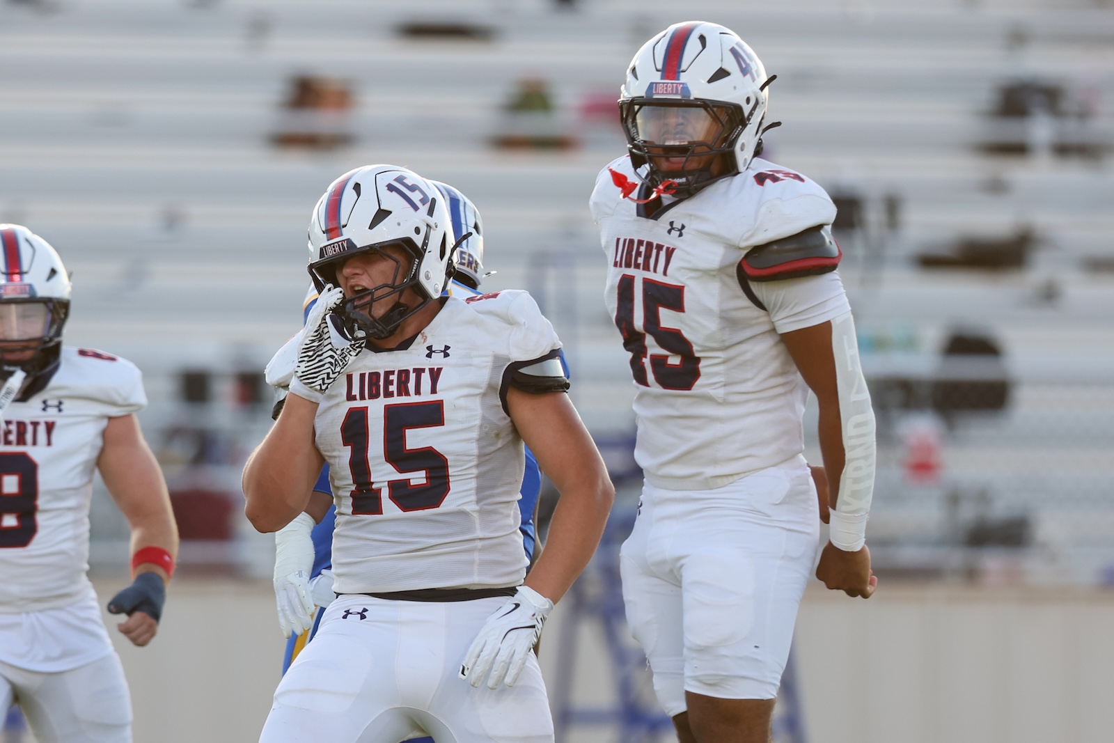 Seabolt Chase-17193 | Press Pass Sports Liberty football players in white uniforms on the field; foreground player #15 adjusting his helmet with another teammate nearby.