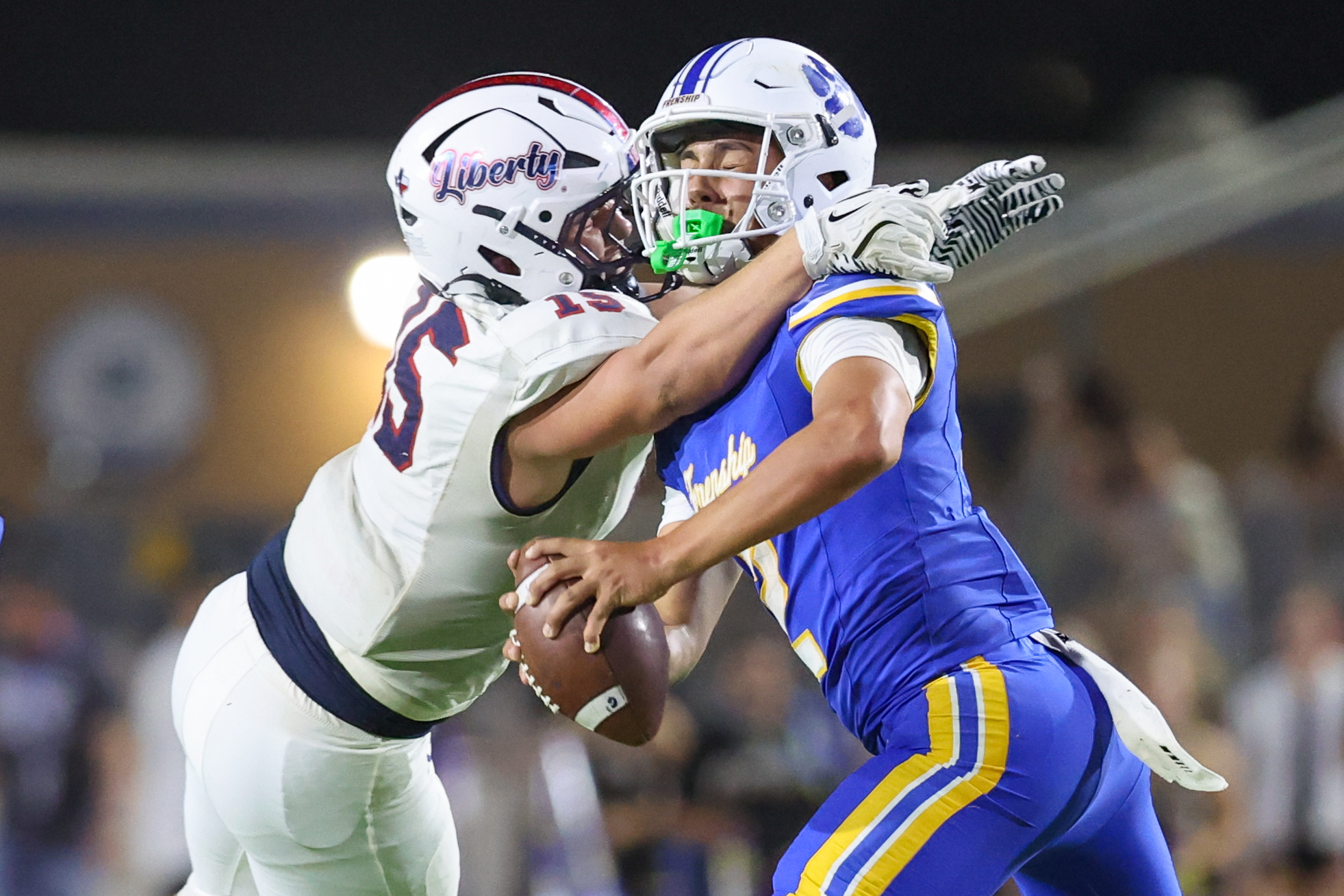 Two football players collide in midair during a play, one in a white uniform with a red Liberty helmet and the other in a blue and yellow uniform, both reaching for the ball.