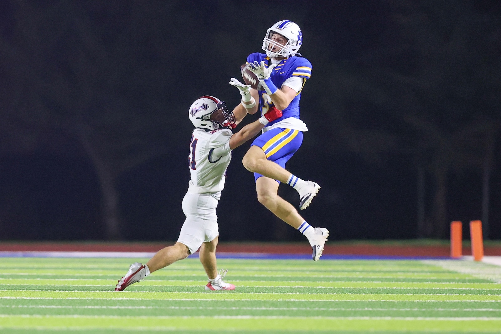 Blue-uniform receiver leaps to catch a pass as a white-uniform defender reaches up during a night football game on a bright turf field.