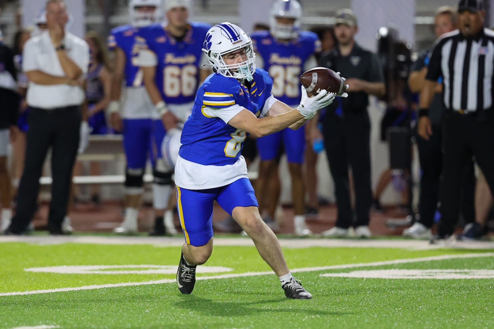 Football player in a blue and white uniform makes a catch on bright green turf with teammates and a referee in the background,