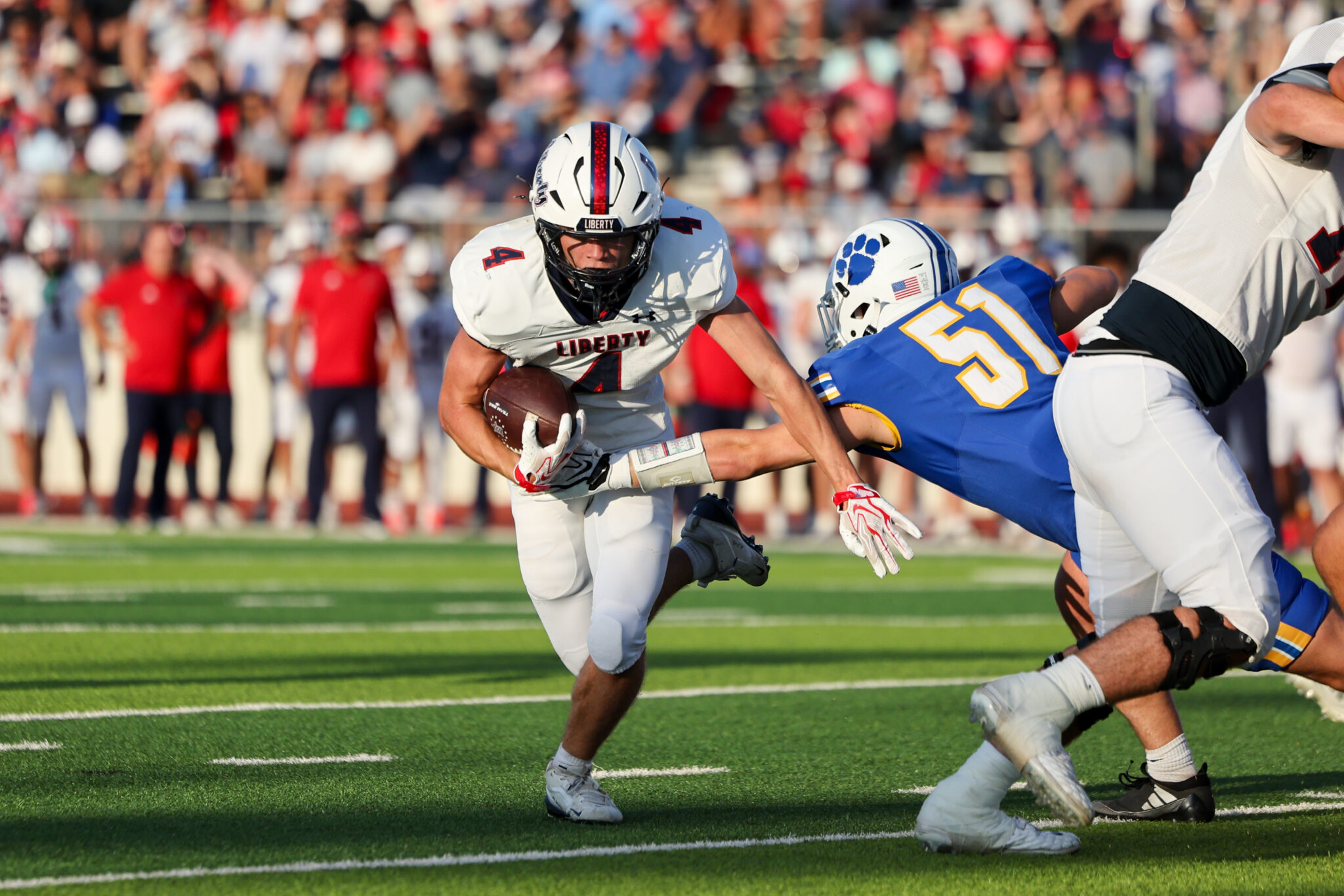 College football: a player in white Liberty jersey runs with the ball while defenders close in on a sunny field with a crowded stands behind.