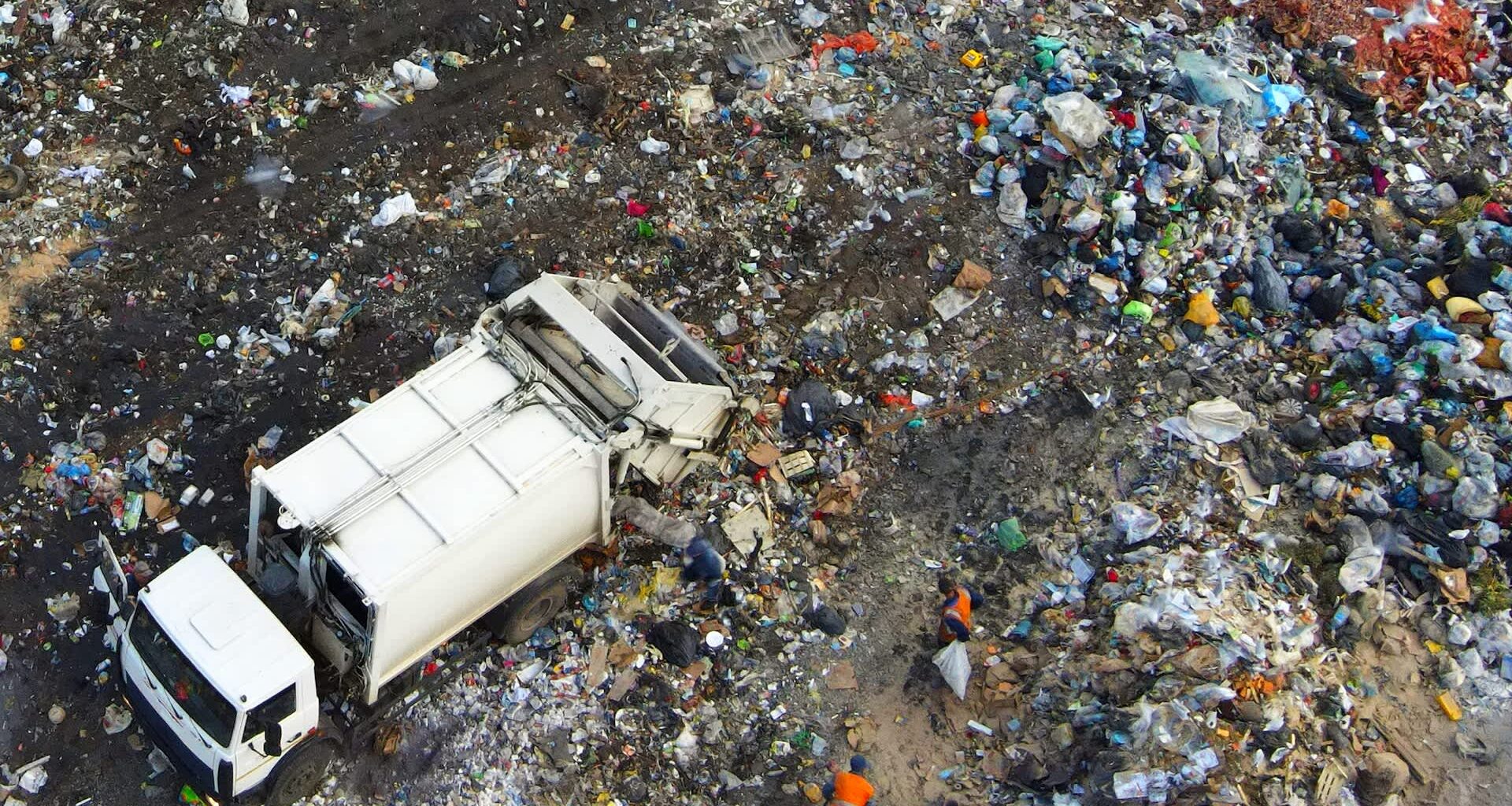 A garbage truck is on a waste pile, with workers collecting debris in a landfill area.