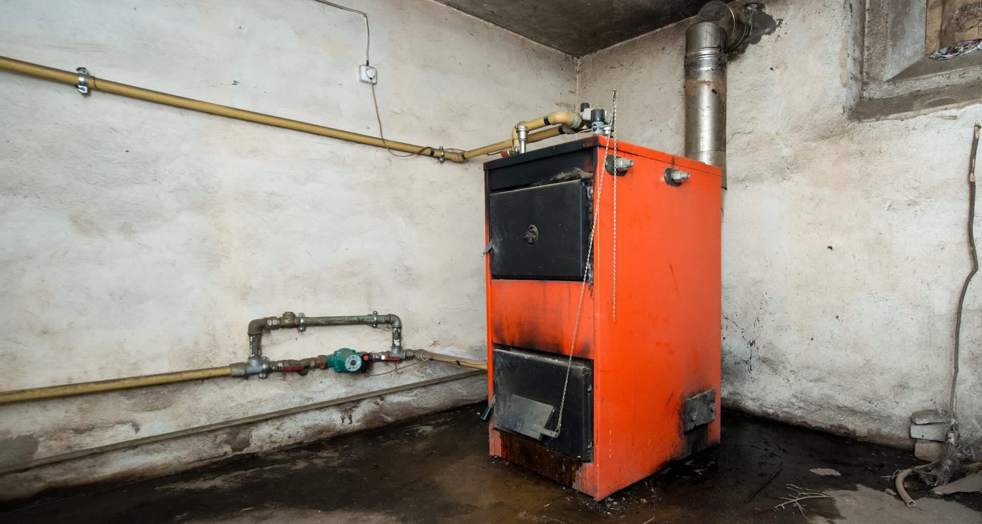 An orange heating boiler is positioned against a bare wall with visible pipes and a damp floor.