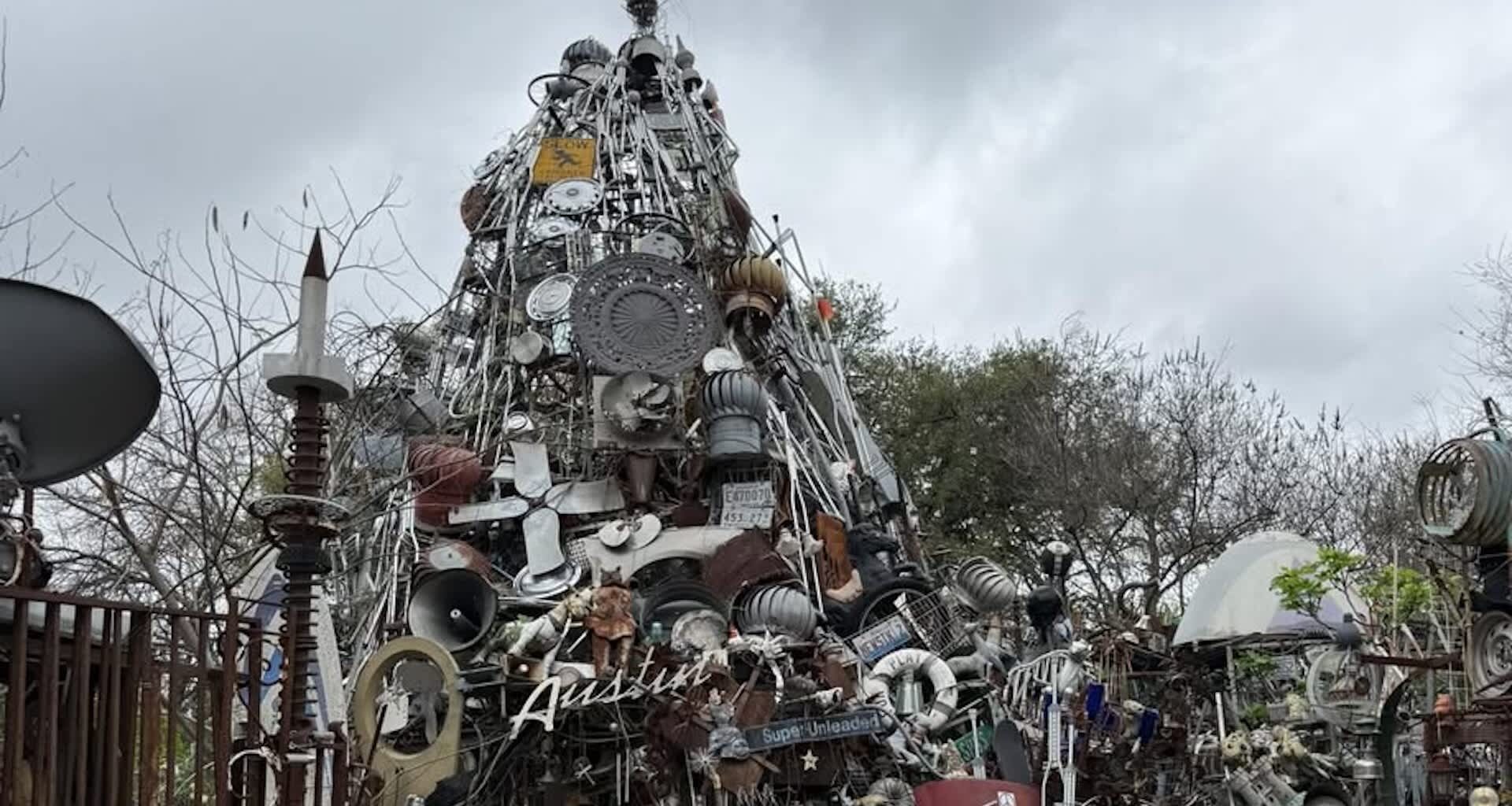 A sculpture made of various metal objects and materials, forming a tower-like structure under a cloudy sky.