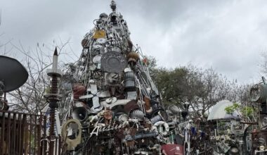 A sculpture made of various metal objects and materials, forming a tower-like structure under a cloudy sky.