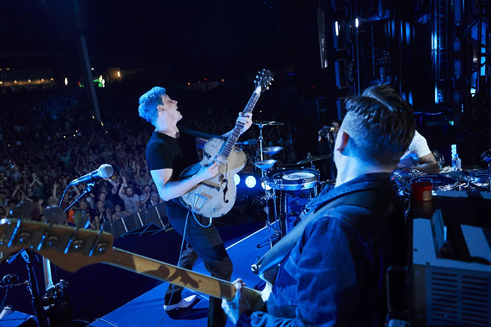 jack white performs on stage with blue hair and wearing all black front of a large crowd while holding a white guitar
