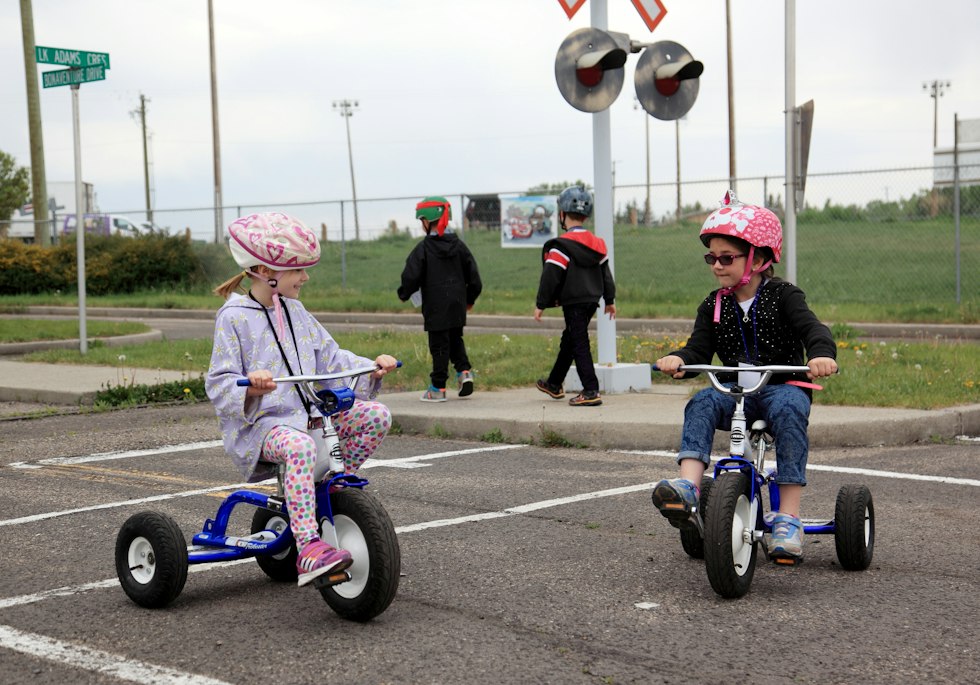 Two children wearing helmets ride tricycles on a paved course