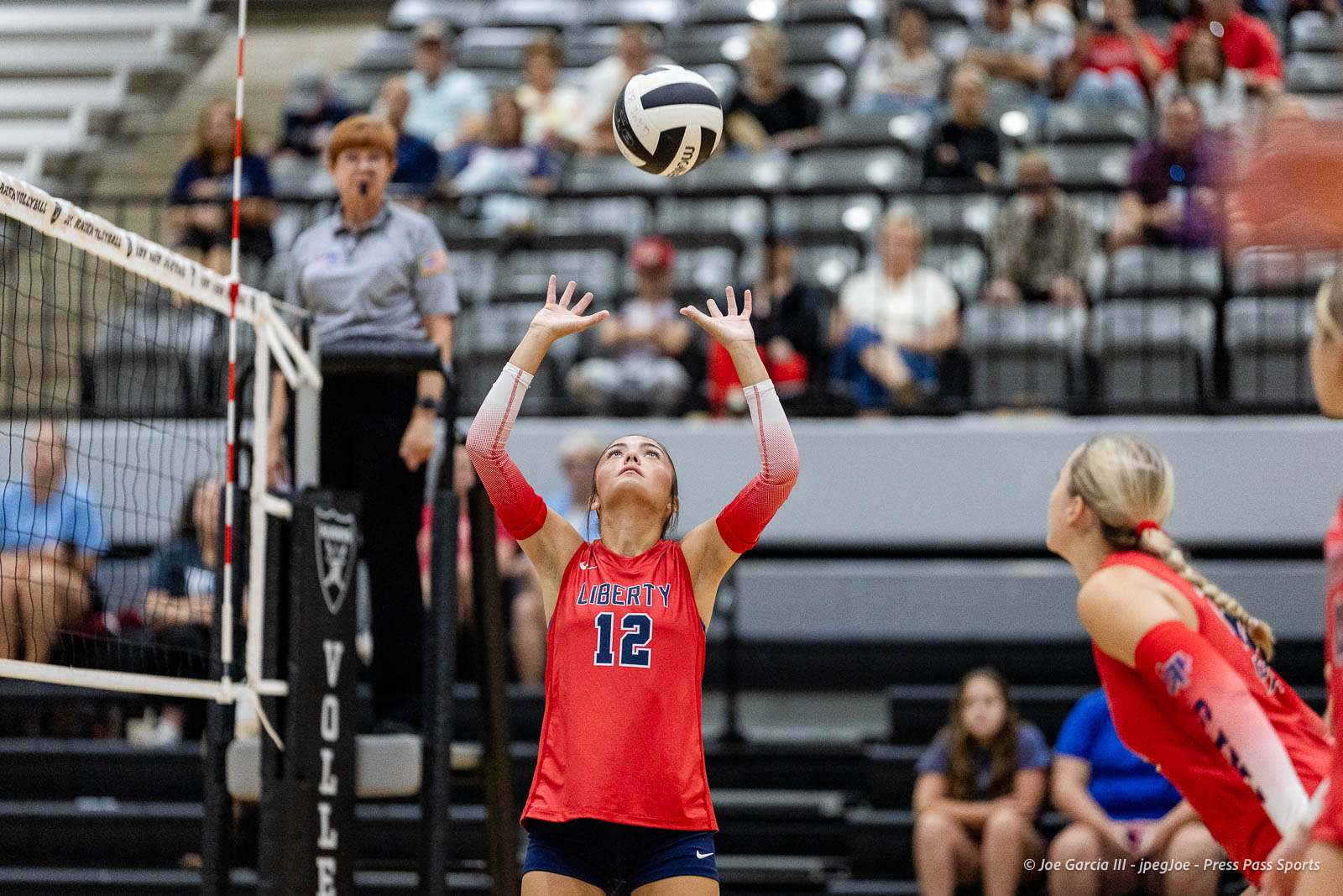 Liberty player #12 in red uniform raises hands to set the volleyball during a rally, with a referee and audience in the background.