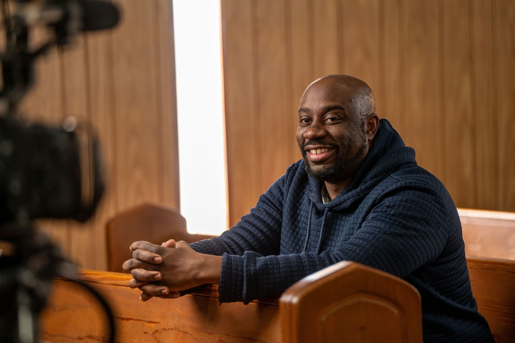 Derek Avery smiles while seated indoors in front of a wood-paneled wall, with a video camera visible in the foreground.