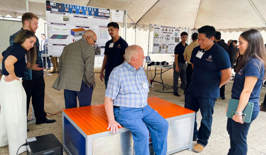 Visitors gather at a student engineering expo while a man tests an orange cooling bench, surrounded by posters and engaged presenters.