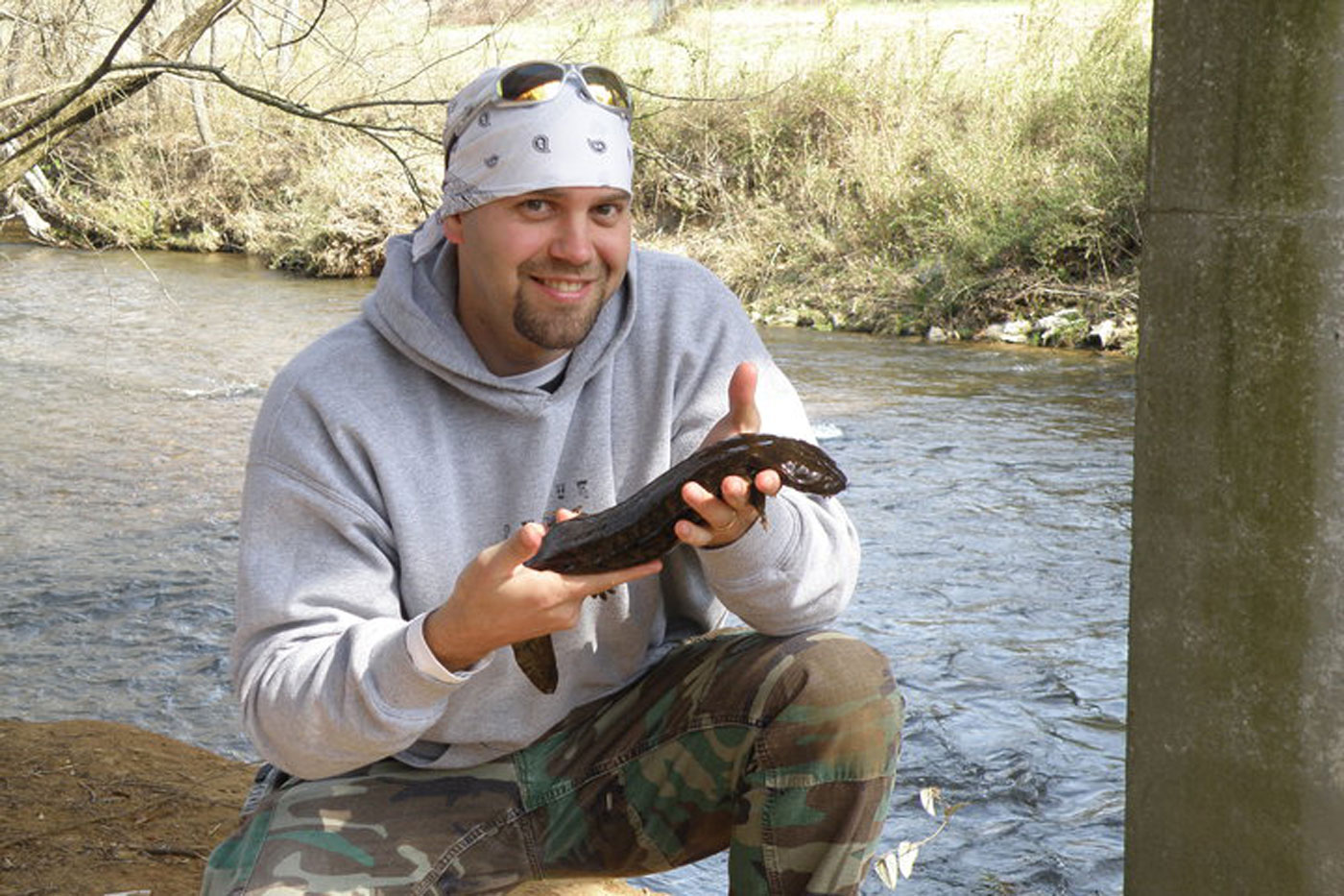 Williams smiles while crouching along the edge of a stream, holding a large hellbender salamander with both hands during field research. Wearing a gray hoodie, camouflage pants and a white bandana, the researcher is surrounded by flowing water and wooded creek habitat.
