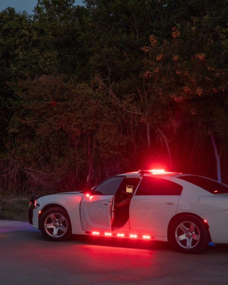 A white Charger police vehicle at night, pulled over by the roadside, with red lights on the car illuminated and the driver’s door cracked open to reveal a screen and someone inside.