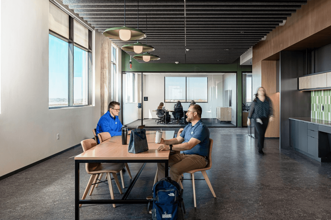 Two people chat at a table in a modern office break area as a colleague walks past a glass-walled meeting room.