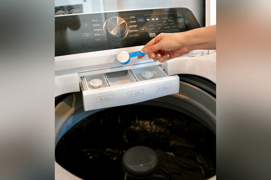 Hand adding powdered detergent with a scoop to the dispenser drawer of a top-load washing machine.
