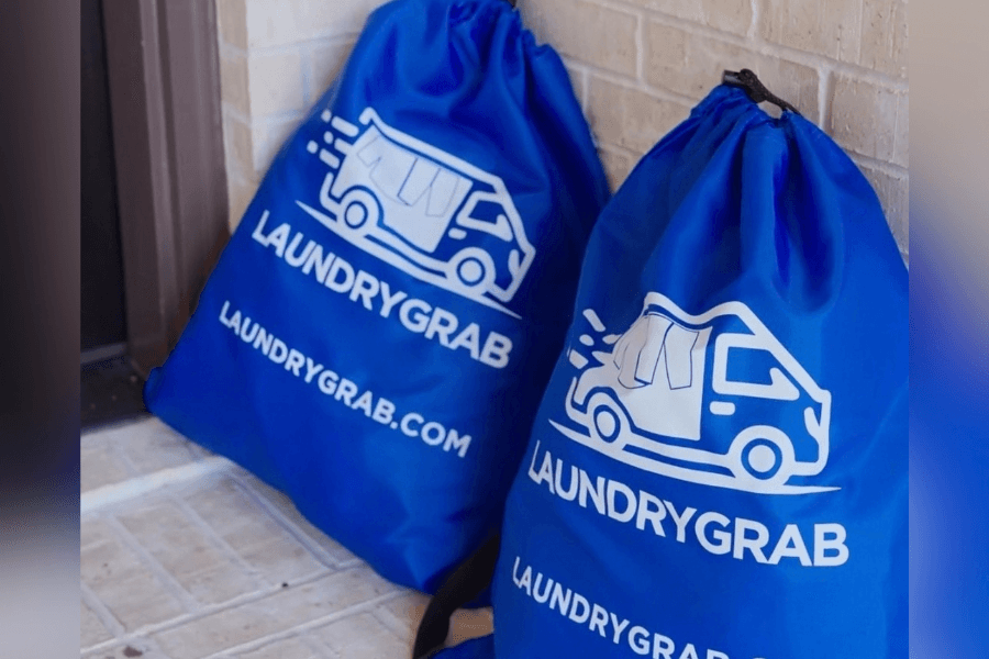 Two blue drawstring laundry bags branded "LaundryGrab" with a truck logo, set against a brick wall.