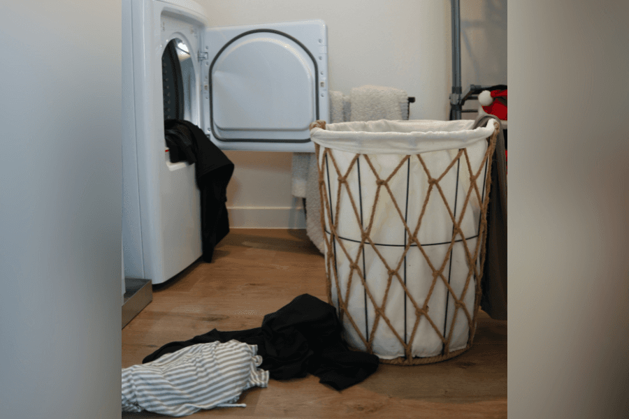 Laundry room with open dryer, rope hamper lined in white, and clothes scattered on wood floor.
