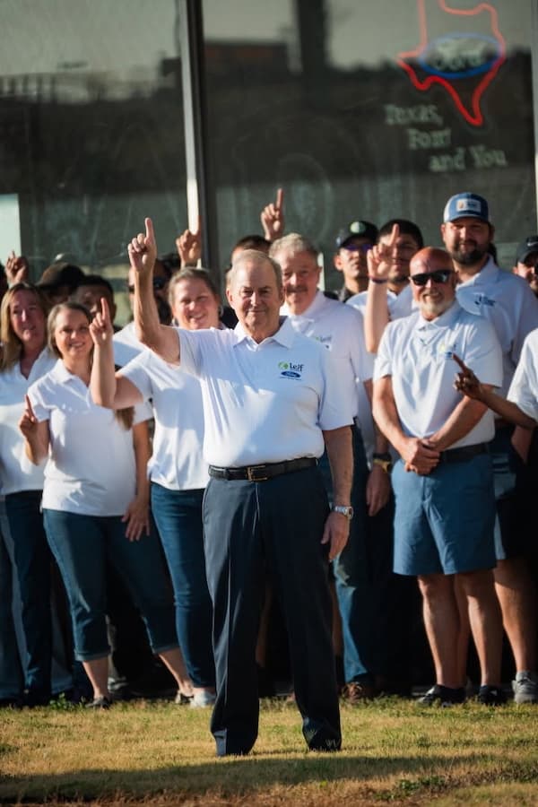 Group in white polo shirts raise index fingers outside a Ford dealership with neon Texas sign.