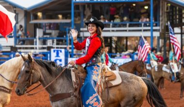 A woman atop a brown horse wears a black cowgirl hat, bright red shirt, and blue chaps and smiles and waves at the camera