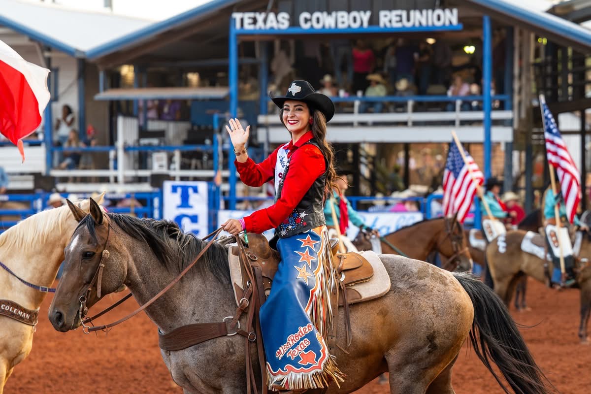 A woman atop a brown horse wears a black cowgirl hat, bright red shirt, and blue chaps and smiles and waves at the camera