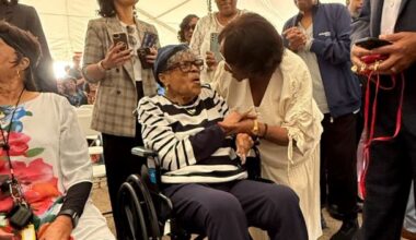 Wheelchair-using elderly woman in a striped shirt shakes hands with a standing woman at a crowded social gathering.