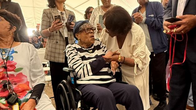 Wheelchair-using elderly woman in a striped shirt shakes hands with a standing woman at a crowded social gathering.